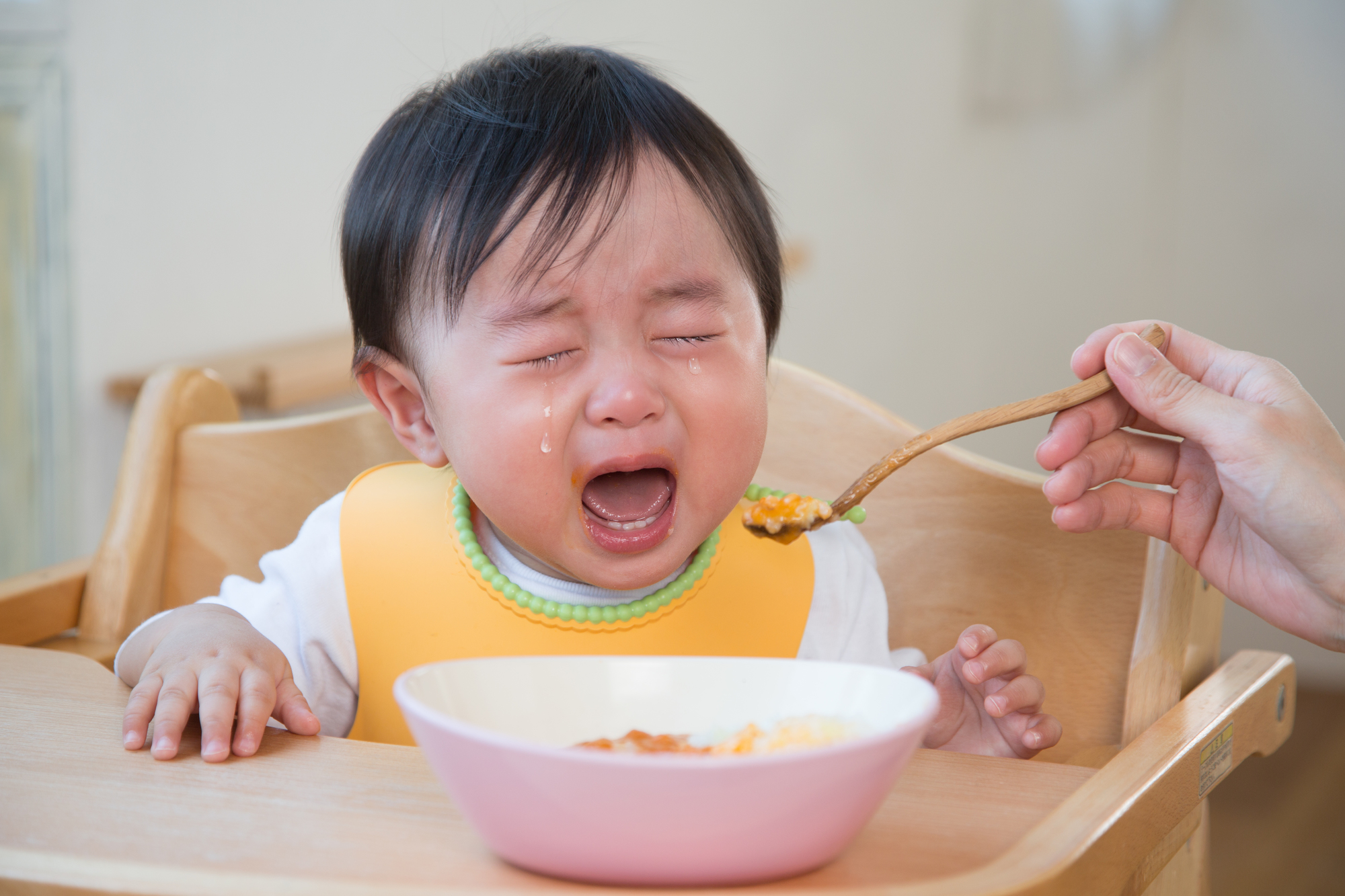 離乳食を食べずに泣く赤ちゃん。ハイチェアでスプーンを嫌がる様子